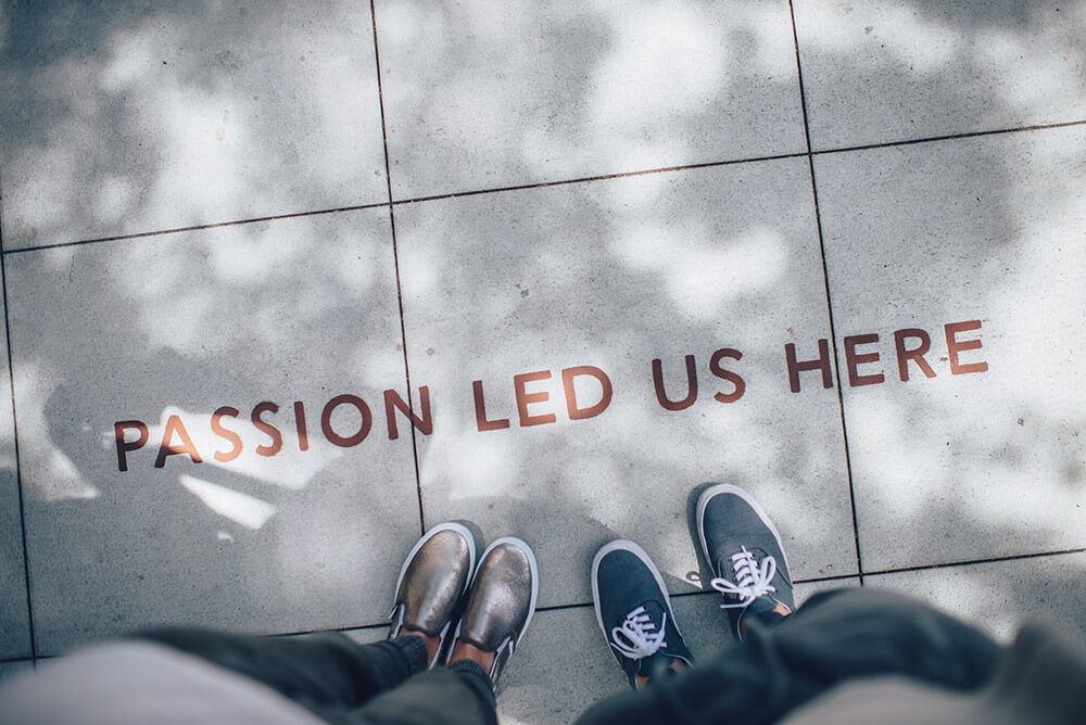 Looking down to a cement tiled walkway with the words 'Passion Led Us Here' and two pairs of feet